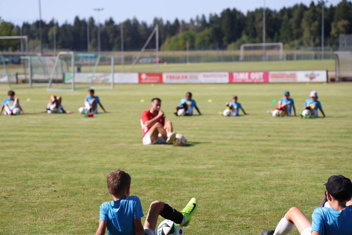 Kinder dehnen sich mit dem Ball auf dem Rasen, der Coach zeigt die Übung vor